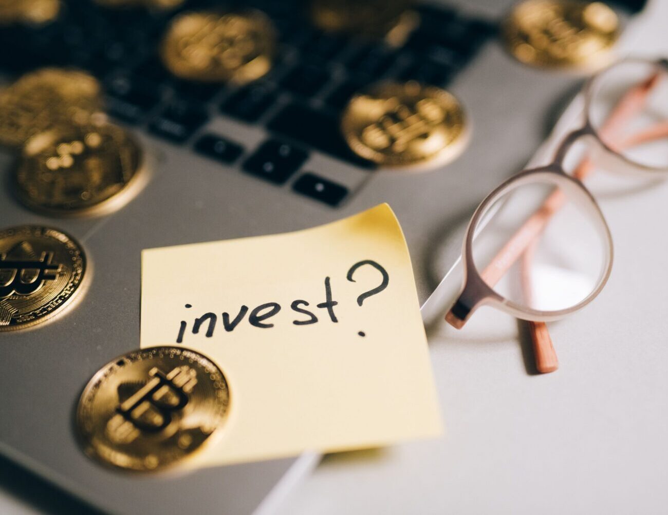 A close-up of bitcoin coins on a laptop with eyeglasses and a sticky note reading 'invest?'